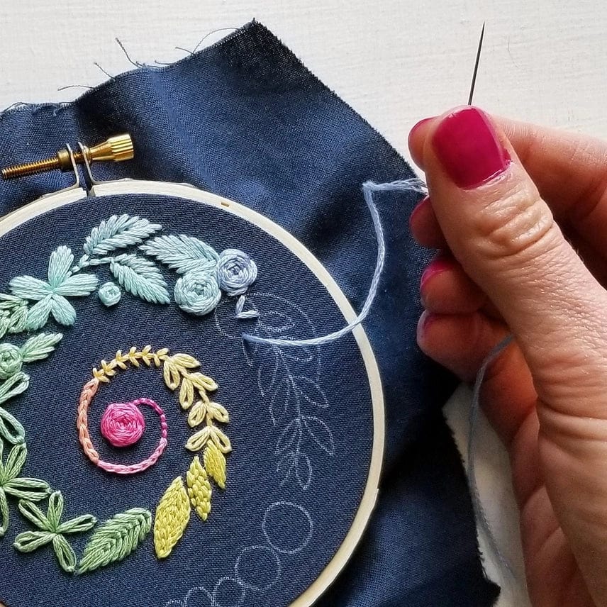 A hand with pink nail polish stitches a colorful floral pattern onto dark blue fabric held in a hoop, using light blue thread and the Jessica Long Embroidery 4 Beginner Navy Spiral Sampler Embroidery Kit—perfect for relaxing.