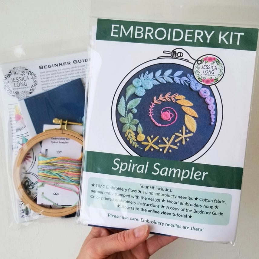 A hand holds the Jessica Long Embroidery 4 Beginner Navy Spiral Sampler Embroidery Kit, which features a floral spiral design and includes a hoop, colored threads, needles, fabric, instructions, and a Beginner Guide booklet.
