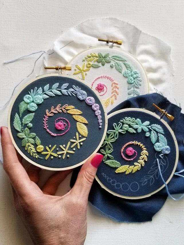 A hand holds the 4 Beginner Navy Spiral Sampler Embroidery Kit by Jessica Long Embroidery, featuring a pastel spiral floral design—ideal for embroidery beginners; two similar hoops nearby highlight this calming hobby on various fabrics.