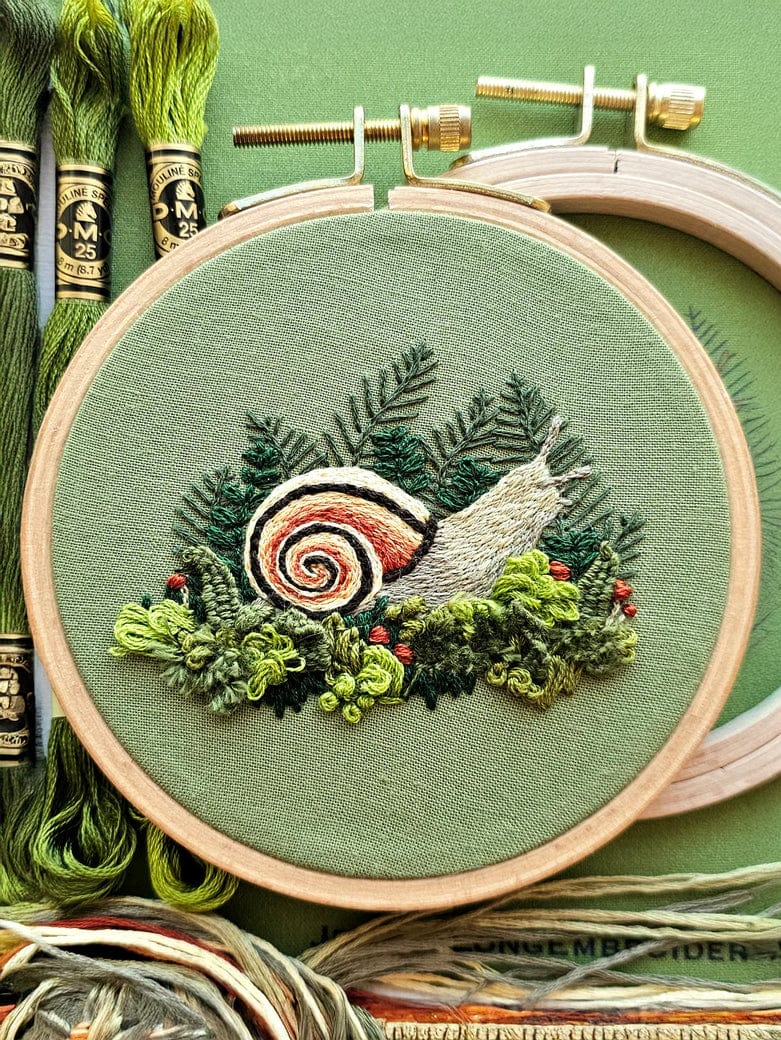The 4 Magic Snail Embroidery Kit by Jessica Long Embroidery features a hand-embroidered snail with a spiral shell among greenery and red berries—ideal for beginners or as thread painting inspiration. Includes green floss on a matching background.