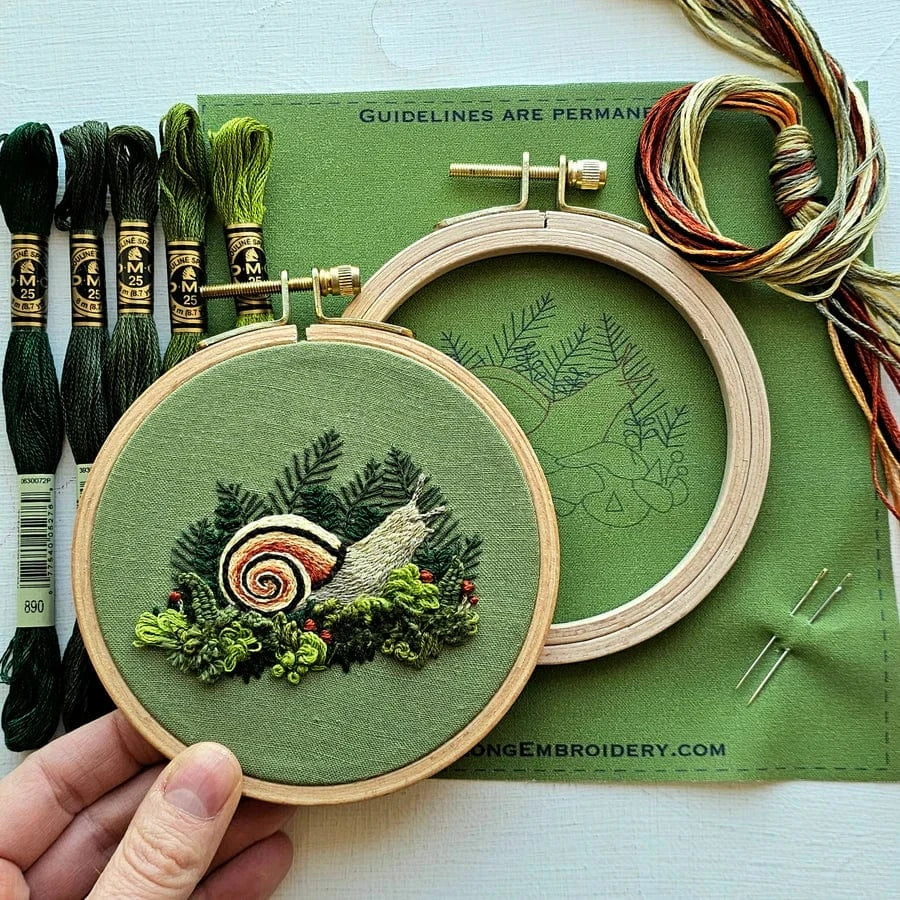A hand holds Jessica Long Embroidery’s 4 Magic Snail Embroidery Kit, featuring a stitched snail on green fabric—ideal for beginners. Also shown: green and multicolored floss, an outlined hoop, and a needle on fabric.