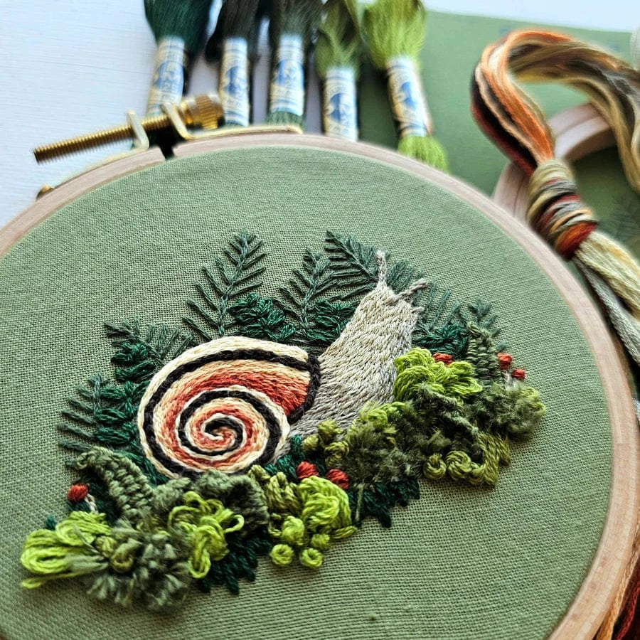 The 4 Magic Snail Embroidery Kit by Jessica Long Embroidery features an embroidered snail with a spiral shell among green plants on fabric in a hoop—ideal for beginners or those exploring thread painting, with colorful threads included.