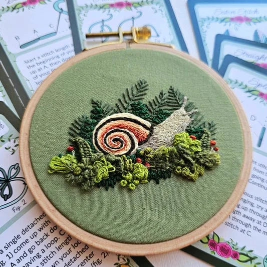 The 4 Magic Snail Embroidery Kit by Jessica Long Embroidery features a snail stitched on green fabric with ferns, moss, and red berries in a wooden hoop. Beginner-friendly instructional cards are partially visible in the background.
