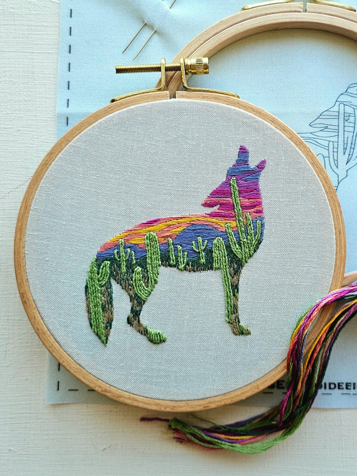 The 5 Beginner Desert Coyote Embroidery Kit by Jessica Long Embroidery features a coyote silhouette filled with vibrant desert landscape embroidery—sunset sky and green cacti—with threads in purple, orange, red, and green. Perfect for beginners.