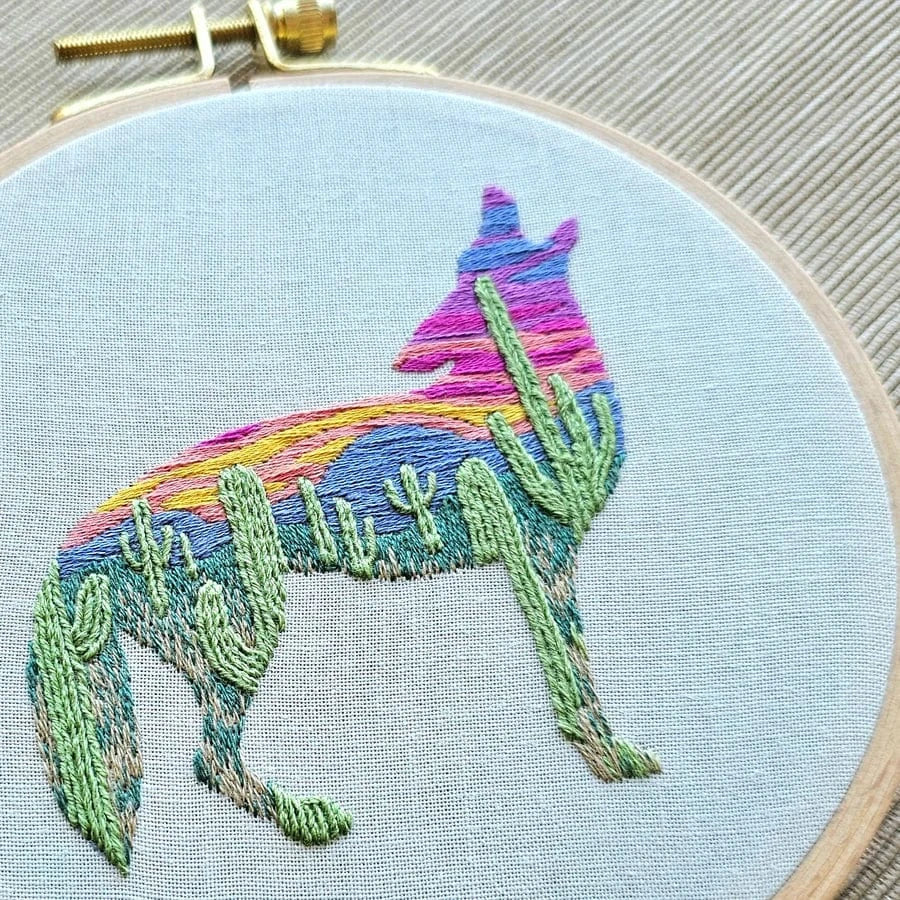 The 5 Beginner Desert Coyote Embroidery Kit by Jessica Long Embroidery features a coyote silhouette with a desert scene—green cacti and a colorful sunset—perfect for beginners, and comes in a circular wooden hoop.