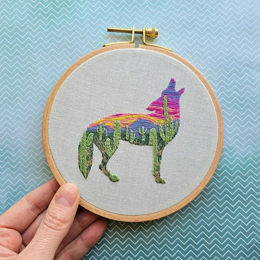 A hand holds the Jessica Long Embroidery 5 Beginner Desert Coyote Embroidery Kit, featuring a coyote silhouette filled with green cacti and a vibrant pink, orange, and purple desert sunset, stitched on light fabric.