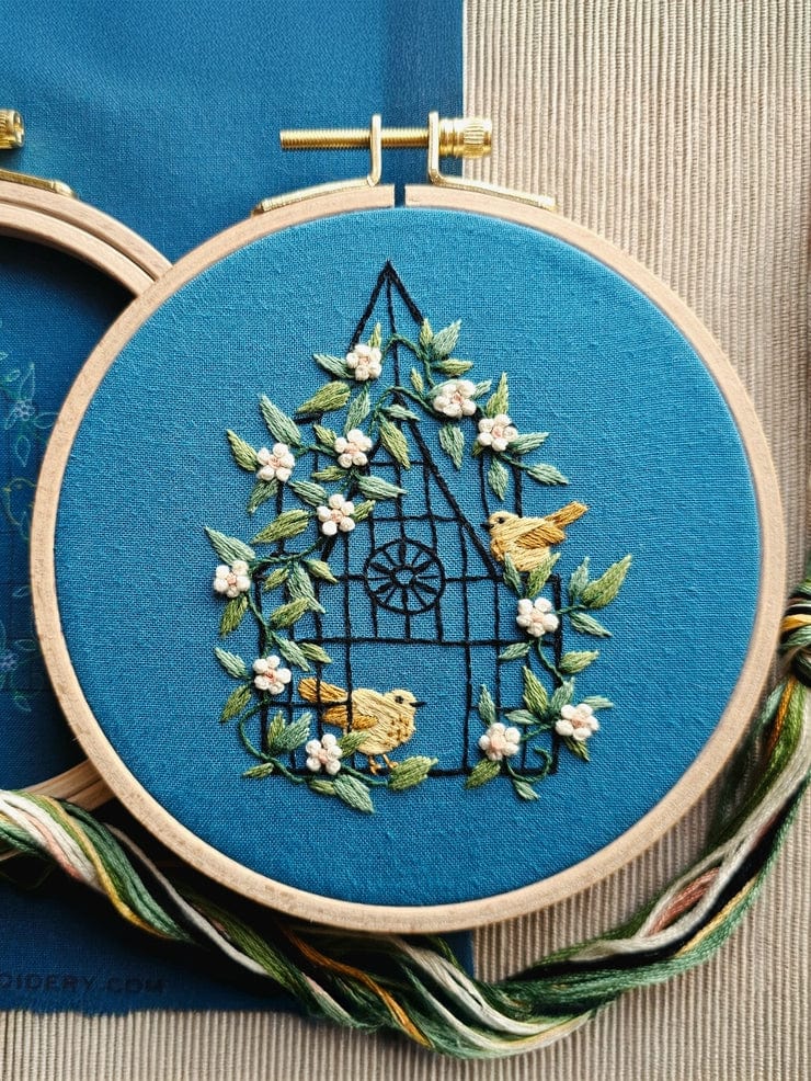 The 5 Beginner Fly Free Embroidery Kit by Jessica Long Embroidery features two yellow birds on a floral wreath with green leaves and white flowers, stitched on bright blue fabric. Embroidery supplies are included beside the hoop.