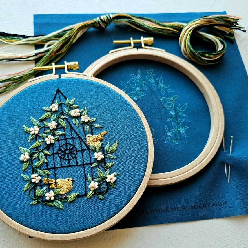 The Jessica Long Embroidery 5 Beginner Fly Free Kit features a blue fabric with birds, white flowers, and green leaves around a window. Included are threads, two needles, and a second hoop with the pattern outline.