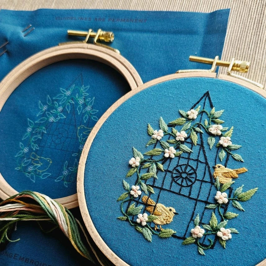 The 5 Beginner Fly Free Embroidery Kit by Jessica Long Embroidery features a blue fabric in a hoop, displaying a stitched house with green vines, white flowers, and yellow birds—an ideal project for beginners.