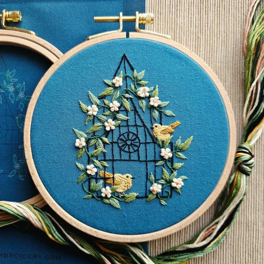 The Jessica Long Embroidery 5 Beginner Fly Free Embroidery Kit features a blue fabric hoop with a black-outlined house, green vines, white flowers, and two yellow birds. Threads are included—ideal for beginner hand embroidery projects.
