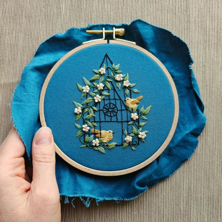 A hand holds Jessica Long Embroiderys 5 Beginner Fly Free Embroidery Kit, showing a birdhouse design with green leaves, pink flowers, and three yellow birds stitched on blue fabric.