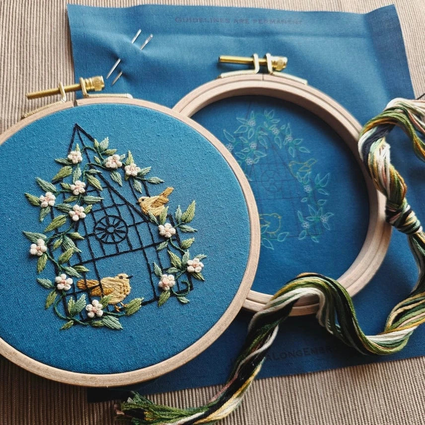 The Jessica Long Embroidery 5 Beginner Fly Free Embroidery Kit features two hoops—one with a finished birdhouse, flowers, and birds, the other with an outline—plus multicolored floss and needles for beginners.