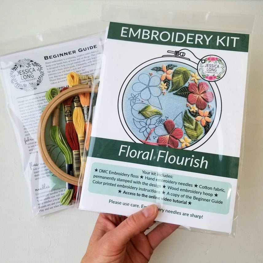 A hand holds the Jessica Long Embroidery *Late May Pre-order* 5 Beginner Green Floral Flourish Embroidery Kit, featuring colored floss, a wooden hoop, a printed floral design, beginner guide, and essentials for floral hoop art.