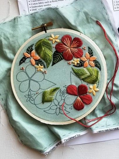 The *Late May Pre-order* 5 Beginner Green Floral Flourish Embroidery Kit by Jessica Long Embroidery includes a light green fabric in a hoop with floral patterns, some brightly stitched in red, yellow, and green thread. A needle with red thread is included.