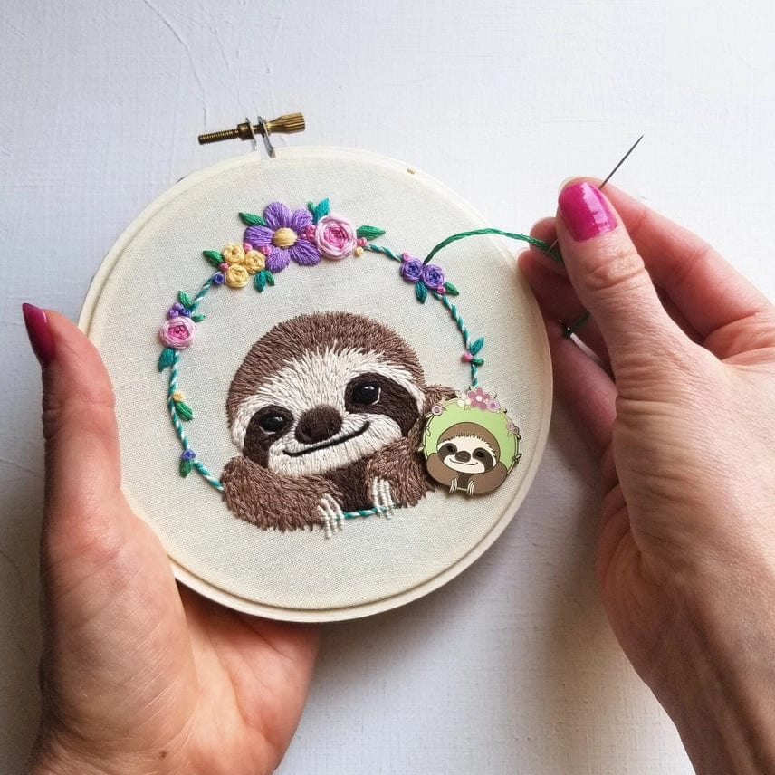 A person embroiders Jessica Long Embroiderys 5 Beginner Smiling Sloth Kit, crafting cute hoop art of a smiling sloth with flowers. Wearing pink nail polish and holding a needle, they’ve added a sloth pin—perfect for handmade DIY gifts.