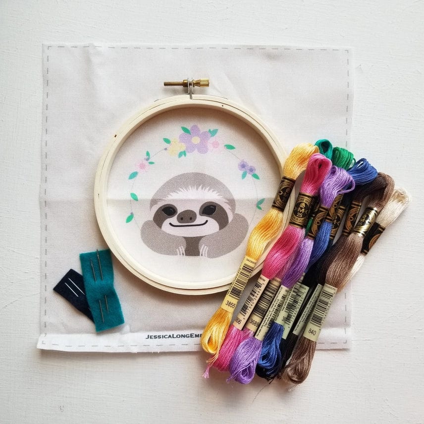 Jessica Long Embroidery’s 5 Beginner Smiling Sloth Embroidery Kit includes a hoop, pre-printed fabric with a cheerful sloth and flowers, colorful floss skeins, two blue felt pieces, and needles—ideal for DIY gifts or hoop art projects.