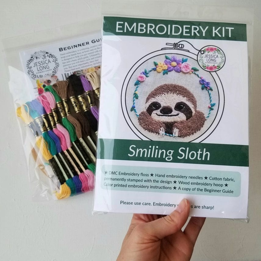 A hand holds the Jessica Long Embroidery 5 Beginner Smiling Sloth Embroidery Kit with a floral sloth design, ideal for DIY gifts or hoop art. Nearby is a clear pack of colorful floss and a Beginner Guide label.