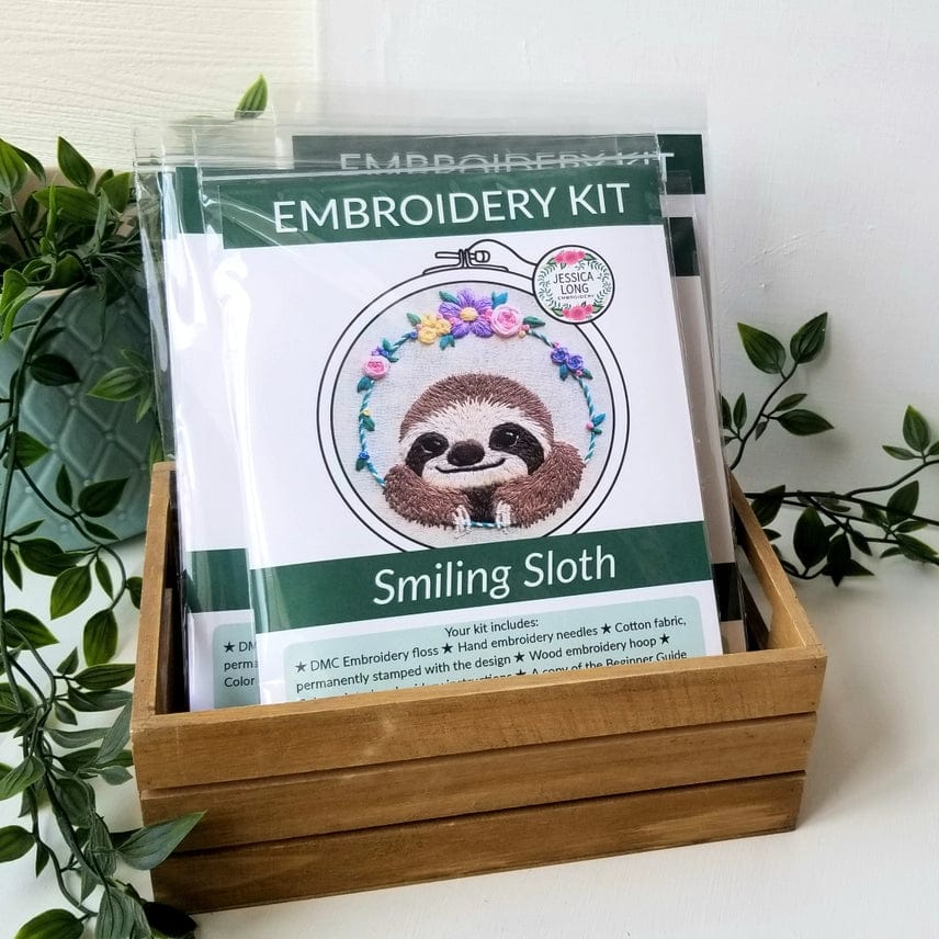 The Jessica Long Embroidery 5 Beginner Smiling Sloth Embroidery Kit is packaged in a wooden box and features a hoop art kit with a smiling sloth and colorful flowers. “EMBROIDERY KIT” and “Smiling Sloth” appear in green on the packaging, perfect for gifting.