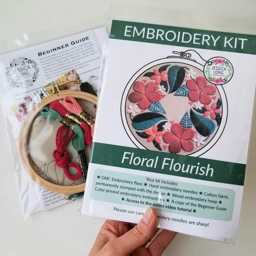 A hand holds the Jessica Long Embroidery 5 Beginner Taupe Floral Flourish Embroidery Kit, featuring a colorful floral pattern, wooden hoop, threads, and beginner guide—all in clear plastic packaging.