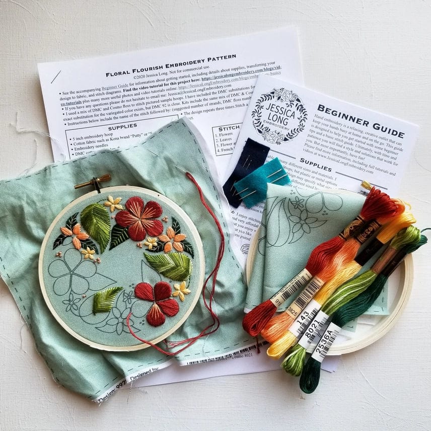 A floral embroidery project in progress on green fabric using the Jessica Long Embroidery 5 Beginner Taupe Floral Flourish Embroidery Kit, with colorful threads, needle, scissors, pattern instructions, and beginner guide arranged on a white surface.