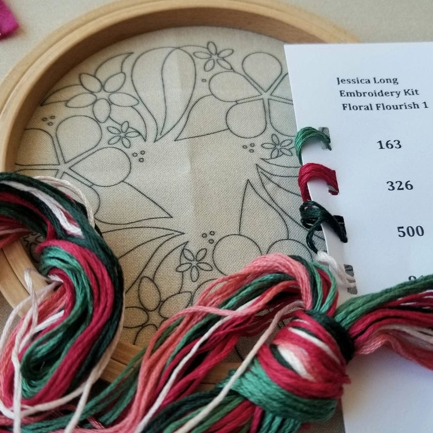 A 5 wooden hoop holds fabric with a floral outline. Green, pink, and white floss bundles sit nearby, along with a card labeled “Jessica Long Embroidery 5 Beginner Taupe Floral Flourish Kit” listing thread numbers.