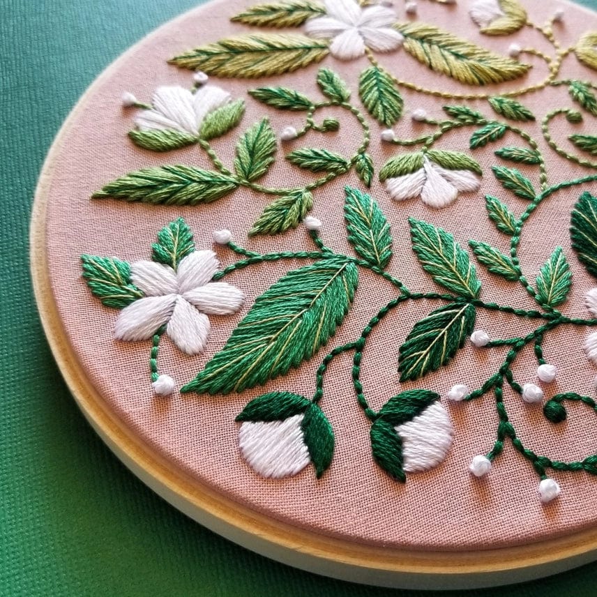 The Jessica Long Embroidery 5 Beginner Blissful Blooms Embroidery Kit features green leaves and white flowers stitched on light brown fabric, showcasing delicate details and thread texture against a dark green background.