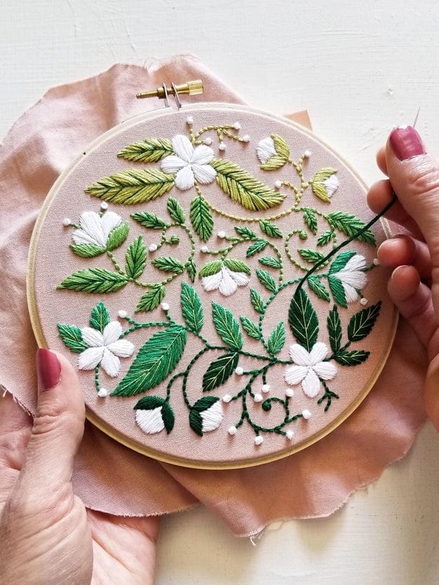 A person uses the 5 Beginner Blissful Blooms Embroidery Kit by Jessica Long Embroidery, stitching green leaves and white flowers with colorful threads—one hand holds the hoop, the other guides the needle. Ideal for a thoughtful handmade gift.