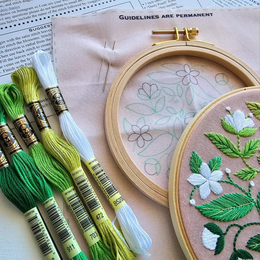 The Jessica Long Embroidery 5 Beginner Blissful Blooms Embroidery Kit includes green and white floss, two needles, a wooden hoop with floral design, finished floral embroidery on pink fabric, and printed instructions—perfect for gifting or beginners.