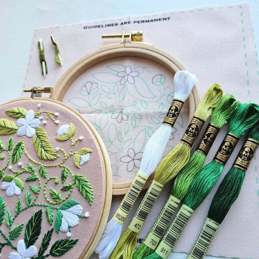 The Jessica Long Embroidery 5 Beginner Blissful Blooms Kit includes a hoop with green and white floral stitching in progress, a second hoop with the pattern, and five matching floss skeins—ideal for crafting a heartfelt handmade gift.