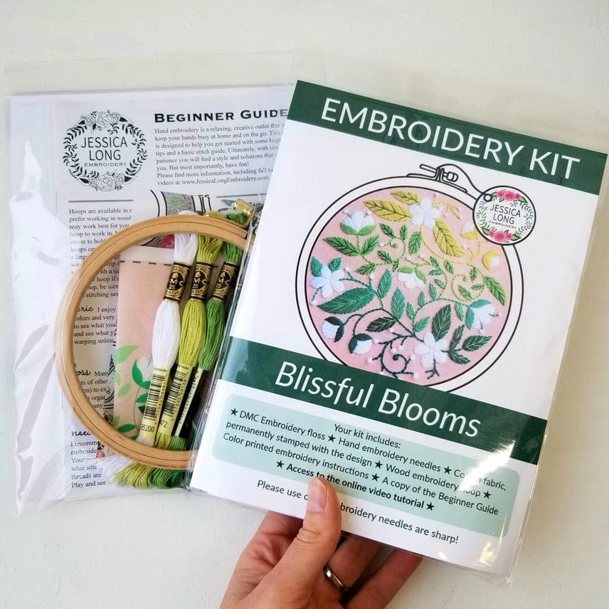 A hand holds the Jessica Long Embroidery 5 Beginner Blissful Blooms Embroidery Kit, featuring a floral pattern, threads, hoop, needle, and beginner guide in clear packaging—a perfect handmade gift for craft lovers.