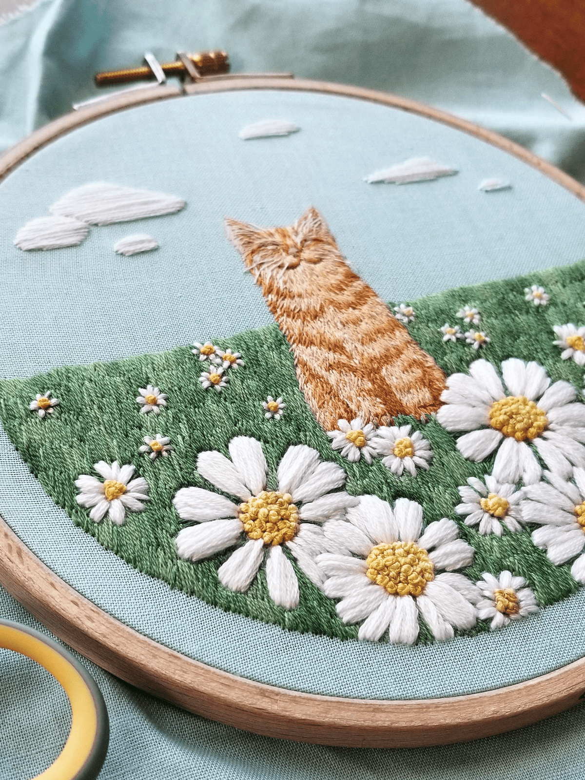 The 5 Happy Cat Embroidery Kit by Jessica Long Embroidery features a stitched orange cat among white daisies on green grass with blue sky and clouds—ideal for beginner embroidery or hand sewing, with a yellow-handled tool included.