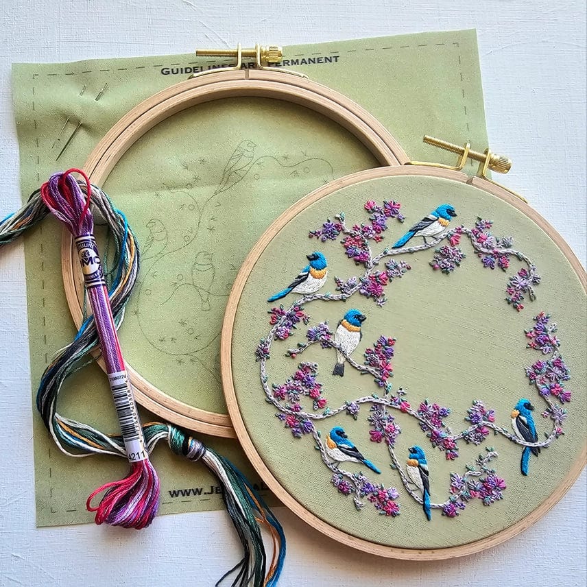 Two 6 hoops from the Jessica Long Embroidery *Late May Pre-Order* Birds & Blooms Kit rest on a flat surface, one with a bird and branch outline, the other featuring stitched blue birds and pink flowers. A bundle of colorful thread lies nearby.