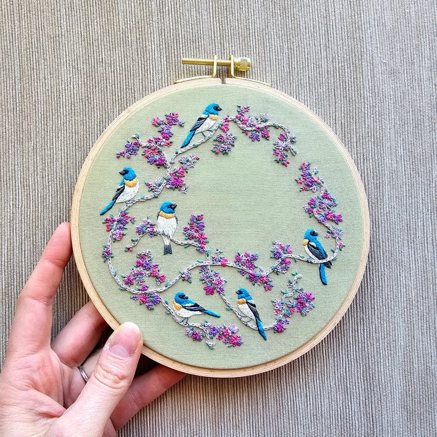 A hand holds the *Late May Pre-Order* 6 Birds & Blooms Embroidery Kit by Jessica Long Embroidery, featuring blue and white birds on branches with pink and purple flowers—ideal for practicing beginner hand embroidery stitches.