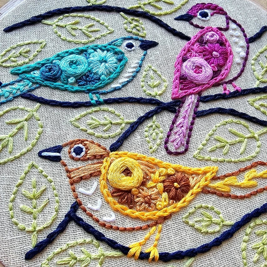 The Jessica Long Embroidery 6 Beginner Colorful Birds Embroidery Kit features three embroidered birds—blue, pink, and yellow—on stitched branches with green leaves, showcasing vibrant colors and intricate textured details on beige fabric.