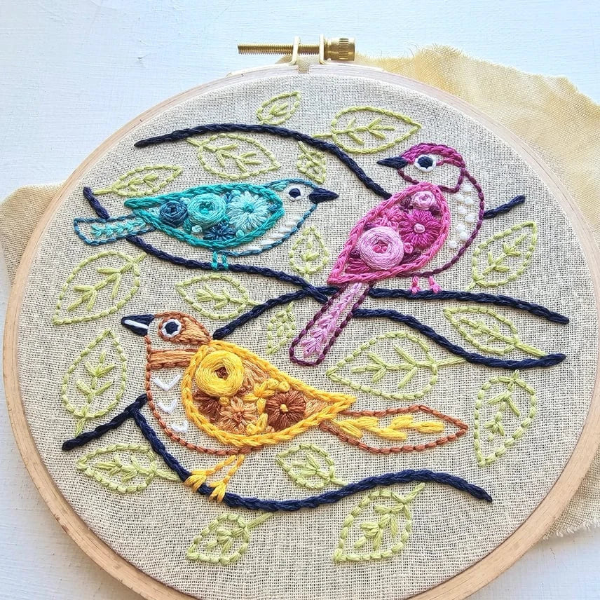 The Jessica Long Embroidery 6 Beginner Colorful Birds Embroidery Kit features blue, pink, and yellow birds on dark branches with green leaves on beige fabric—ideal for beginner hand embroidery projects.