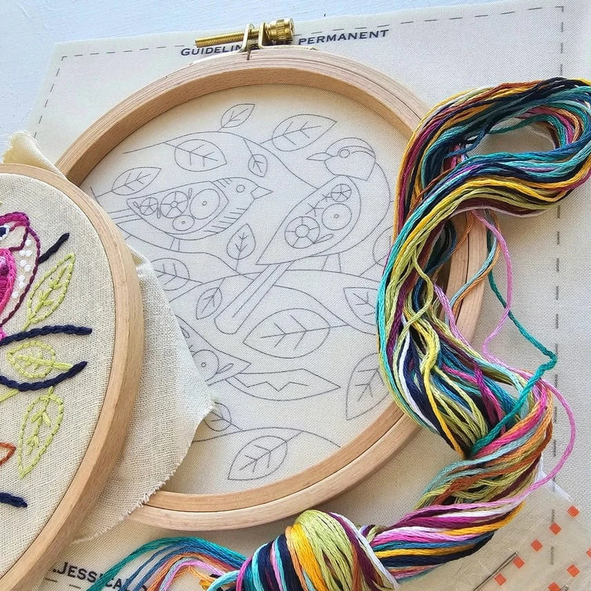 The Jessica Long Embroidery 6 Beginner Colorful Birds Embroidery Kit includes a hoop with outlined bird patterns, a finished colorful bird example, and multicolored threads—perfect for beginners.