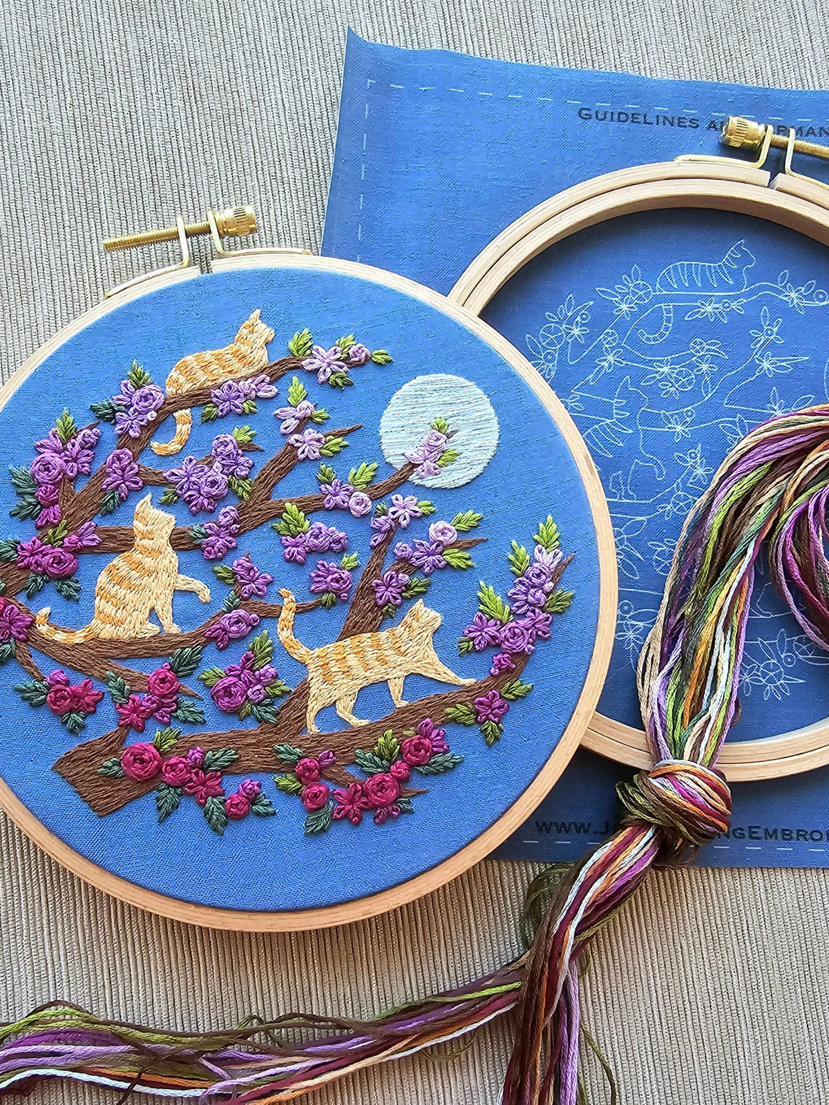 The 6 Beginner Orange Cats & Full Moon Embroidery Kit by Jessica Long Embroidery features three orange cats on a blossoming tree under a full moon, and includes floss, a pattern guide, and easy instructions—perfect for beginners.