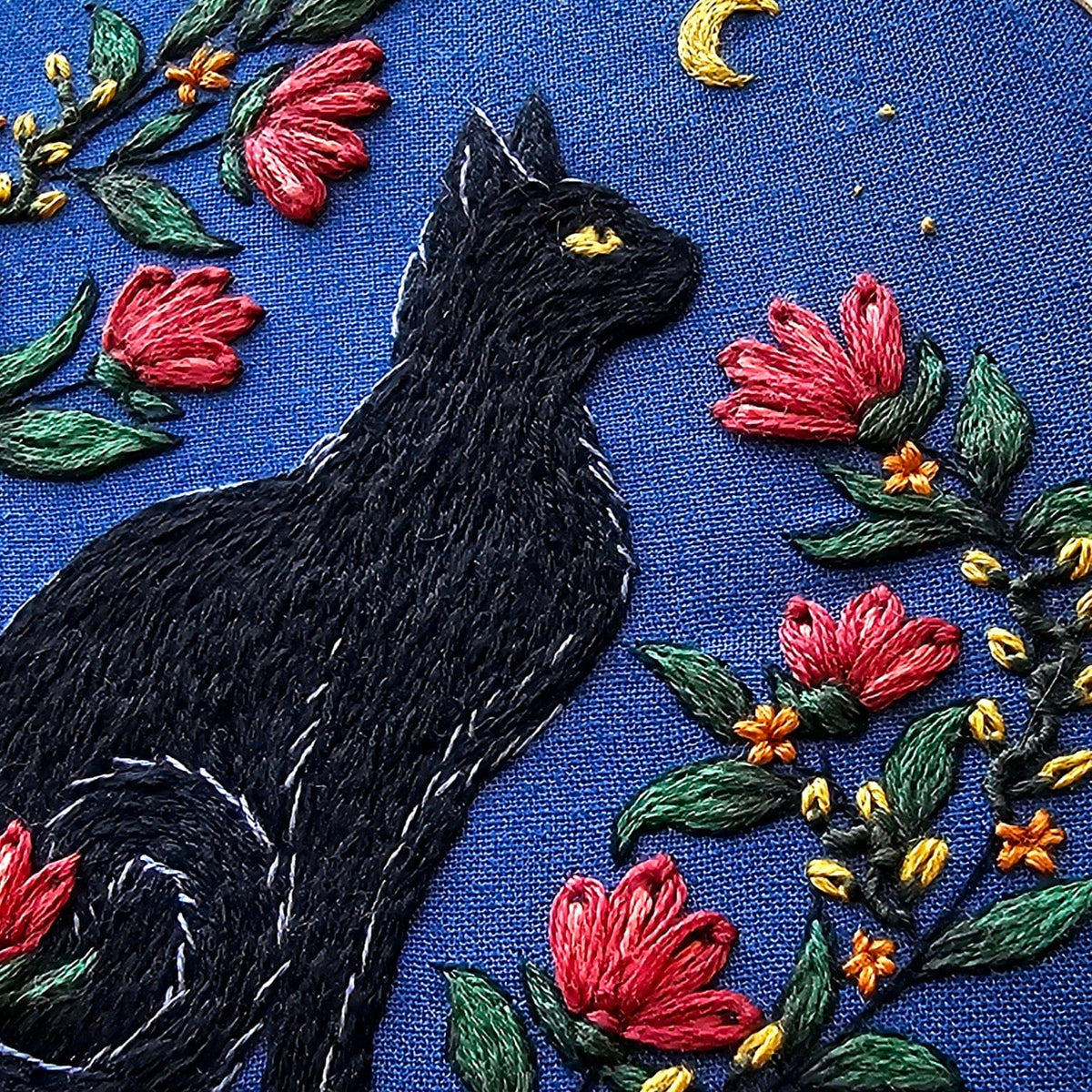 An embroidered black cat gazes at a yellow crescent moon and stars among red flowers on blue fabric in the *Late May Pre-Order* 5 Beginner Stargazer Black Cat Embroidery Kit by Jessica Long Embroidery.