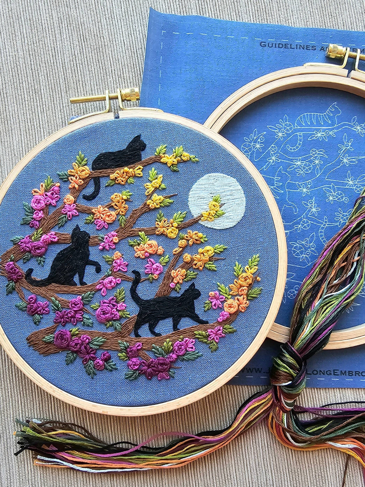 Jessica Long Embroiderys 6 Beginner Black Cats & Full Moon Embroidery Kit features three black cats on a flowering branch under a full moon with vibrant pink, yellow, and orange flowers. Kit includes pattern guide and thread.