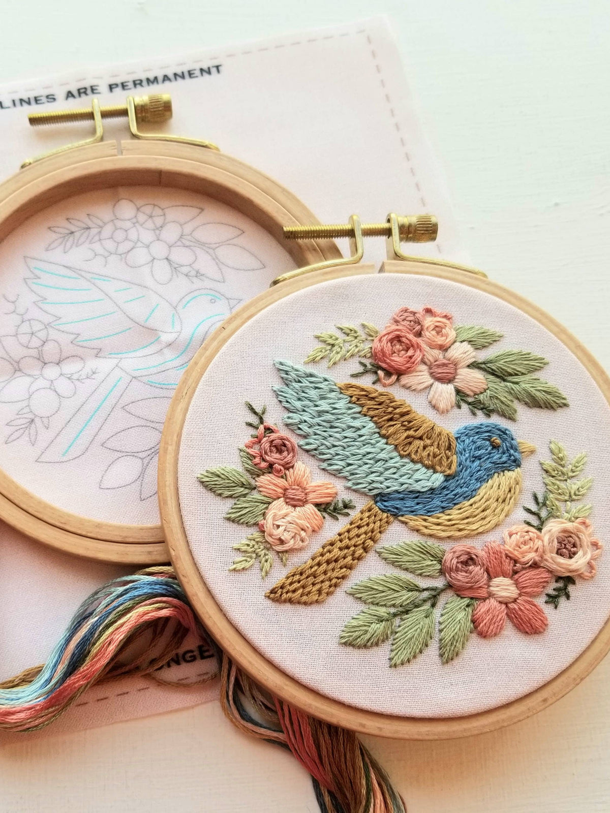 The Jessica Long Embroidery 4 Beginner Bluebird Sampler Embroidery Kit features a hoop holding fabric with a stitched bluebird and flowers, plus an outlined version ready to embroider. Includes visible threads for easy stitching.
