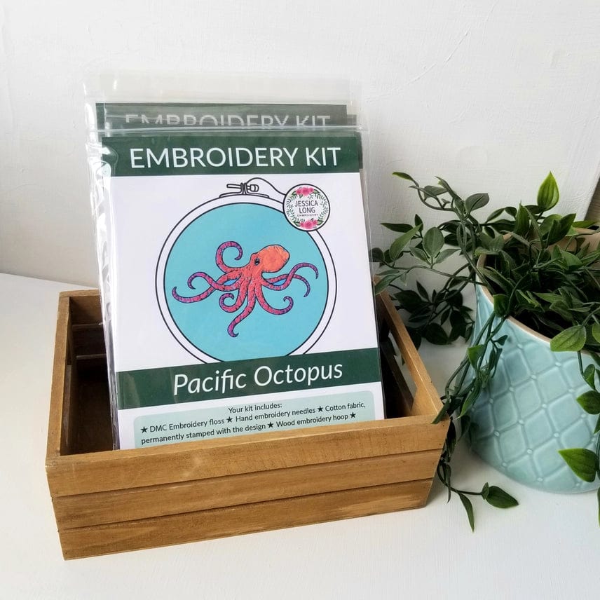 A wooden box holds several Jessica Long Embroidery kits. In front is the 5 Beginner Pacific Octopus Embroidery Kit, featuring a red octopus design. A teal plant pot sits nearby—ideal for ocean wall art enthusiasts.