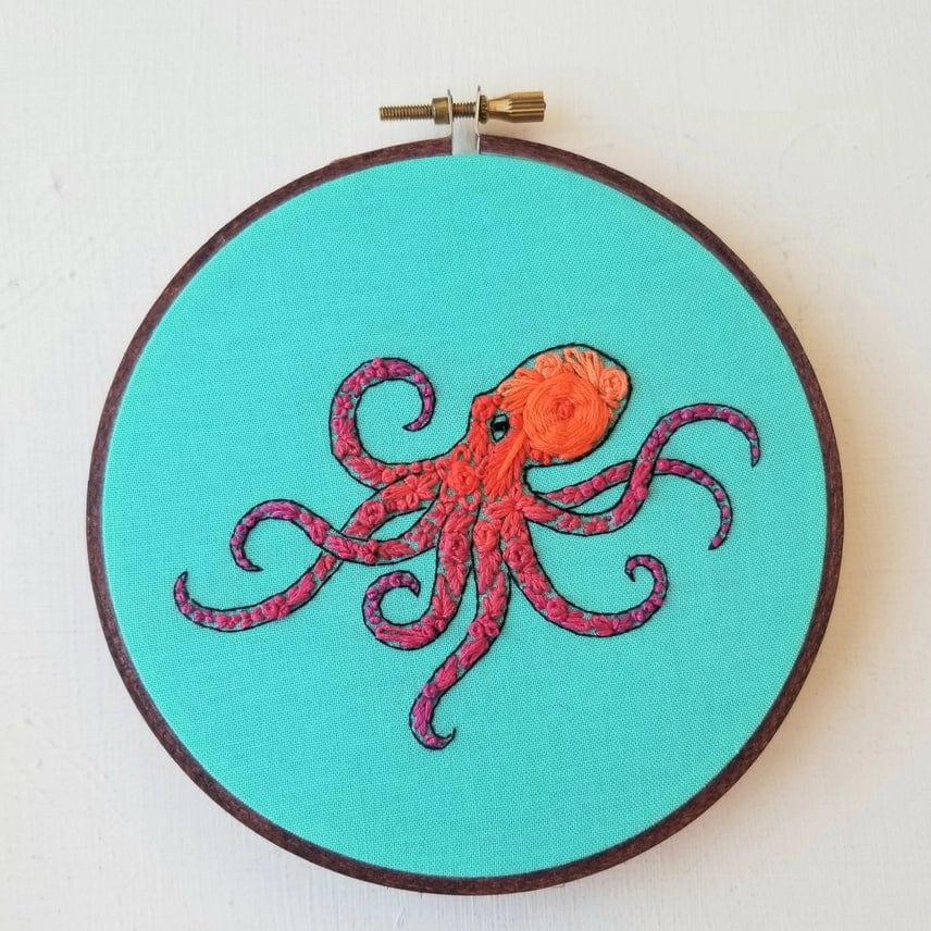 The 5 Beginner Pacific Octopus Embroidery Kit by Jessica Long Embroidery features a red and orange octopus on bright blue fabric in a wooden hoop—a perfect ocean wall art piece or fun sea life craft project.