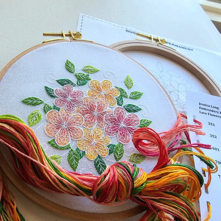 The 6 Lacy Flowers Embroidery Kit by Jessica Long Embroidery features white fabric held in a hoop, colorful stitched flowers and leaves, bright embroidery floss, a pattern guide, and an extra hoop for your next project.