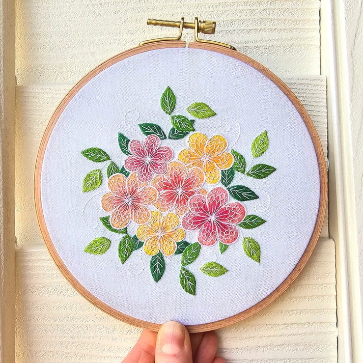 A hand holds the Jessica Long Embroidery 6 Lacy Flowers Embroidery Kit, featuring a vibrant stitched pattern of pink, yellow, and orange flowers with green leaves on a light fabric background.