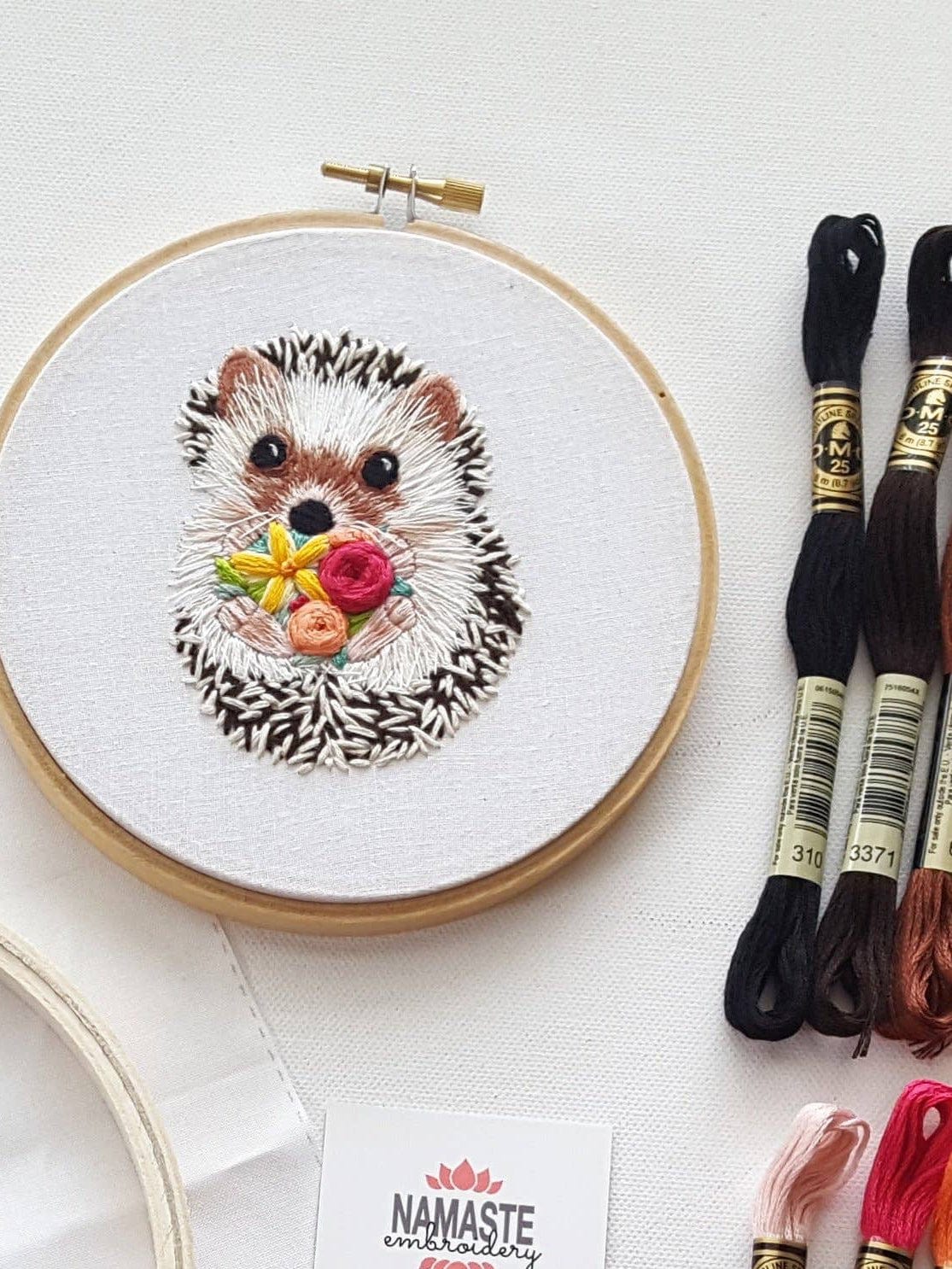 The Jessica Long Embroidery 6 Hedgehog Embroidery Kit features a cute hedgehog holding colorful flowers, with black, brown, and red threads. Great for beginners or as a gift. Namaste Embroidery card included underneath.