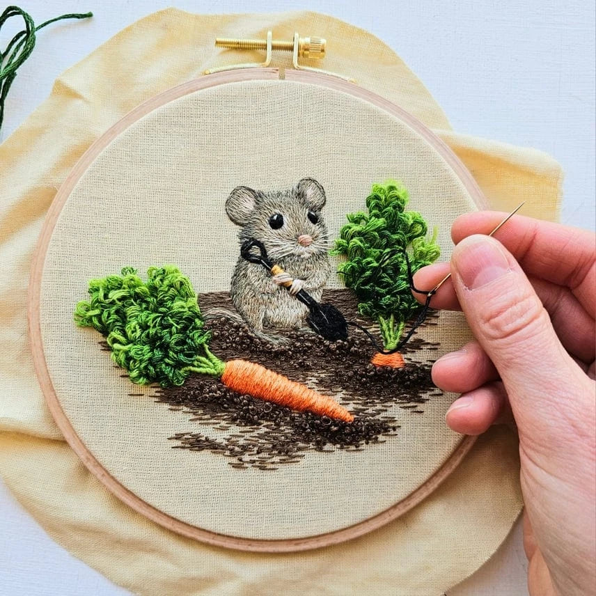 The 6 Garden Mouse Embroidery Kit by Jessica Long Embroidery features a gray mouse with a shovel between two carrots, one being hand-stitched in orange—ideal for thread painting fans. Fabric comes pre-stretched in an embroidery hoop.