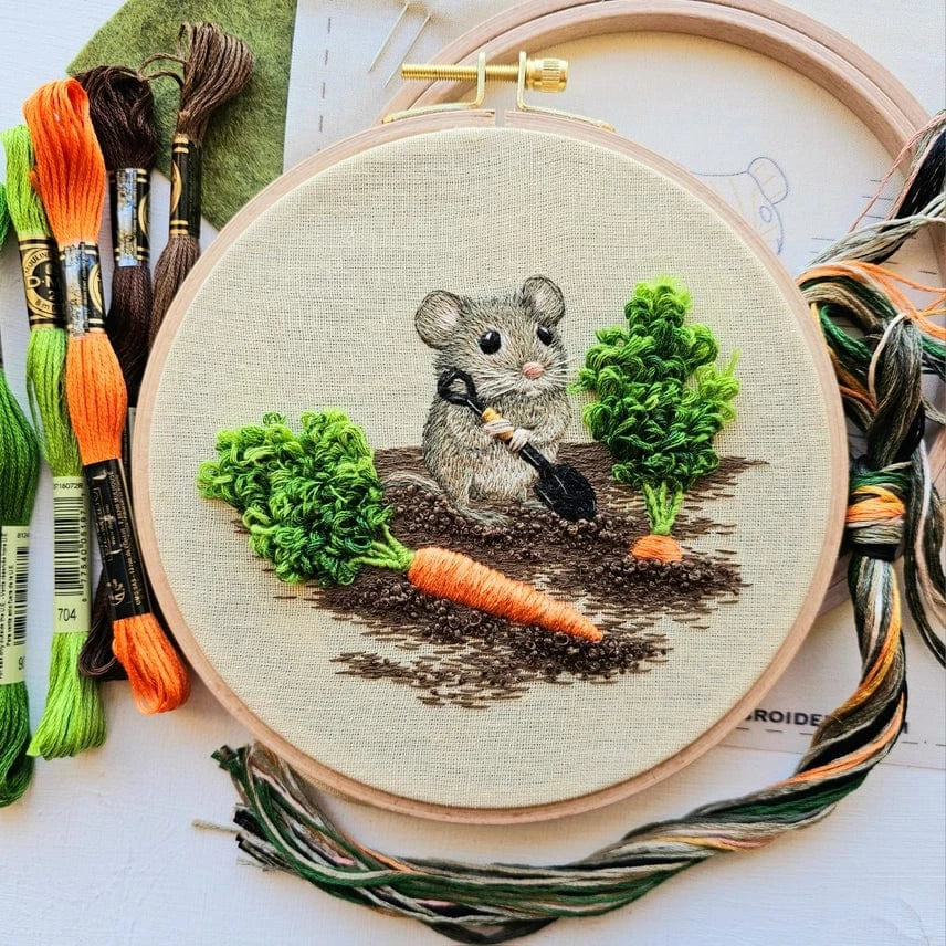 The 6 Garden Mouse Embroidery Kit by Jessica Long Embroidery features a hand-embroidered mouse with a shovel among carrot plants, plus green, orange, and brown threads—perfect for practicing thread painting.