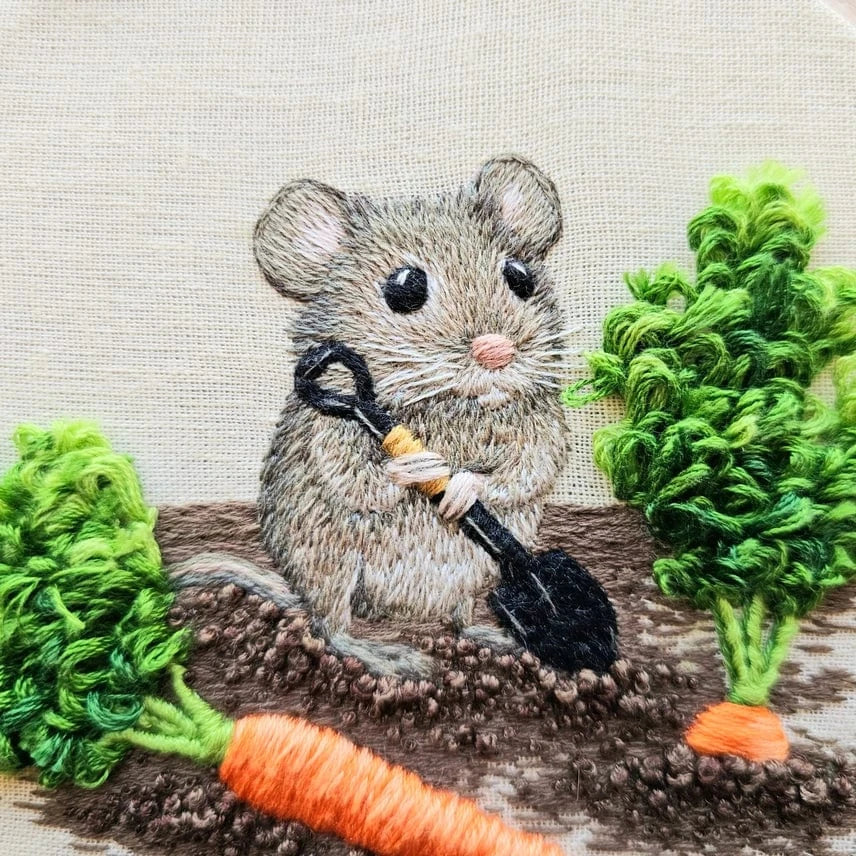 The 6 Garden Mouse Embroidery Kit by Jessica Long Embroidery features an embroidered mouse with a black shovel on brown soil, surrounded by green leafy carrots and one orange carrot below—an adorable stumpwork design for your next project.