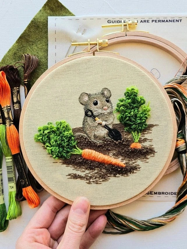 A hand holds the Jessica Long Embroidery 6 Garden Mouse Embroidery Kit, featuring a gray mouse with a shovel digging up a carrot. Colorful threads and the kits pattern are also visible—ideal for thread painting enthusiasts.