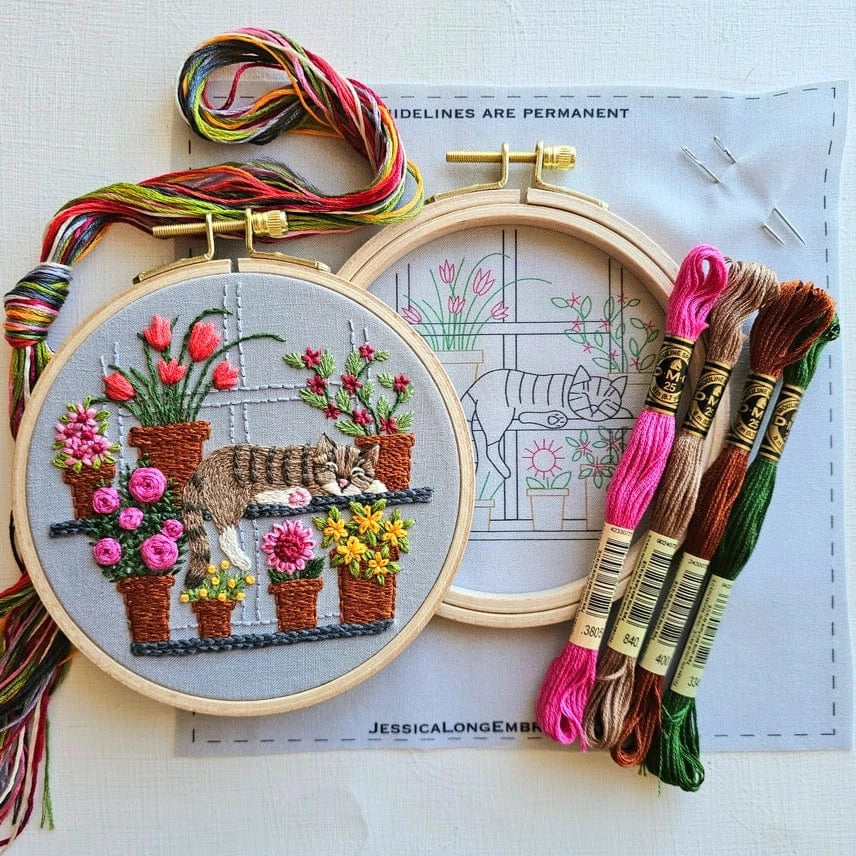The Jessica Long Embroidery 5 Cat Nap with Flowers Embroidery Kit features colorful threads, one finished tabby cat among flowers, and an unfinished hoop on a pattern guide—ideal for beginner floral embroidery projects.